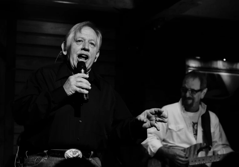 two men standing in a dark room and one talking into a microphone