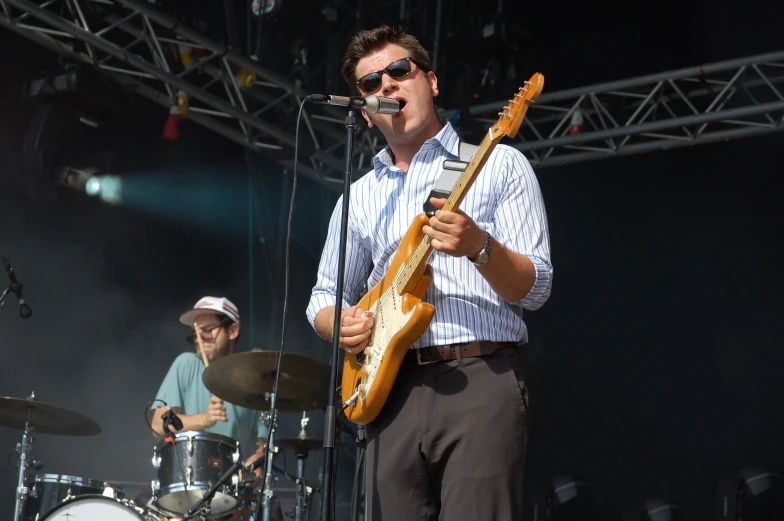 two men singing on stage with guitars, and one holding an electric guitar