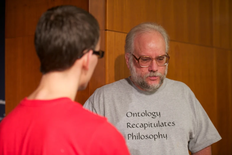 man with greying hair and wearing red standing in front of wall talking to another man