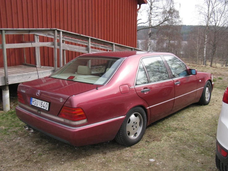 a maroon mercedes cl - type car is parked next to a red barn