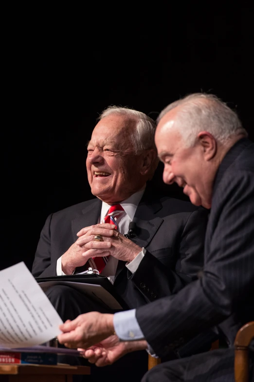 two older men sitting in chairs at a table, one is reading a paper and the other is smiling