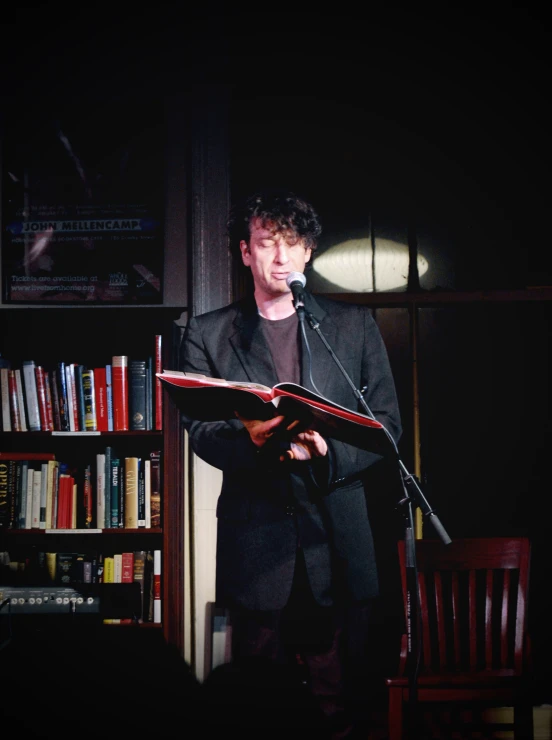man reading from a large open book, with dark bookshelves in the background