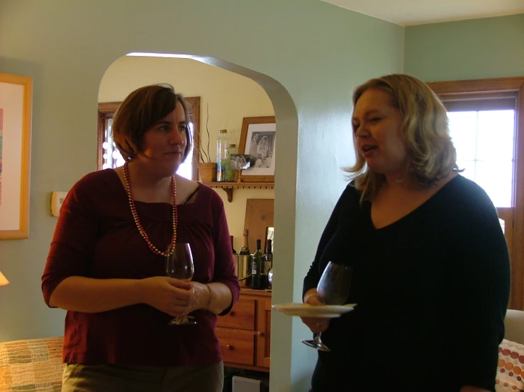 two women standing in a room with one holding a plate and one holding a glass of wine