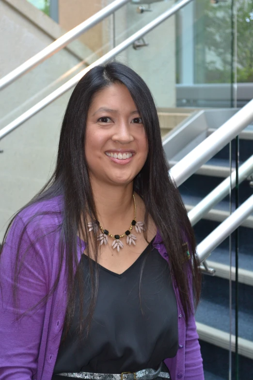 a young woman wearing a black dress and purple jacket stands on some steps smiling