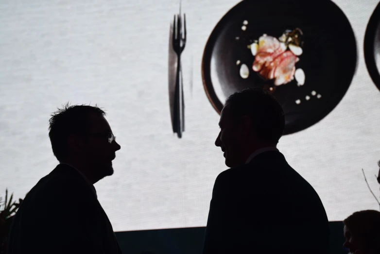 two men talking in front of an event with plates and fork on the screen