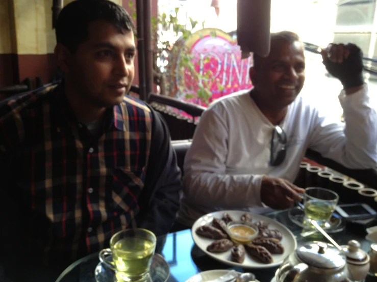 two men are sitting at a table with tea, food and water