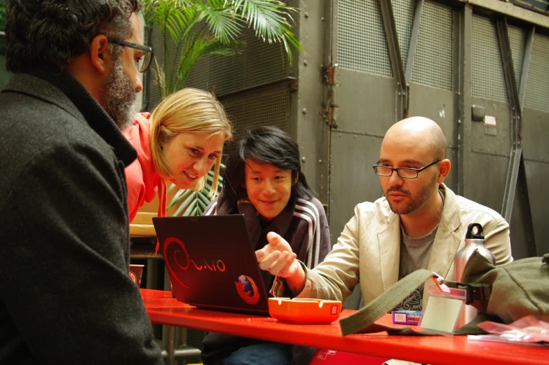 three people sitting at an outdoor table looking at a computer