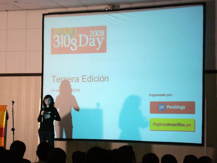 a woman is talking on the stage with a large screen behind her