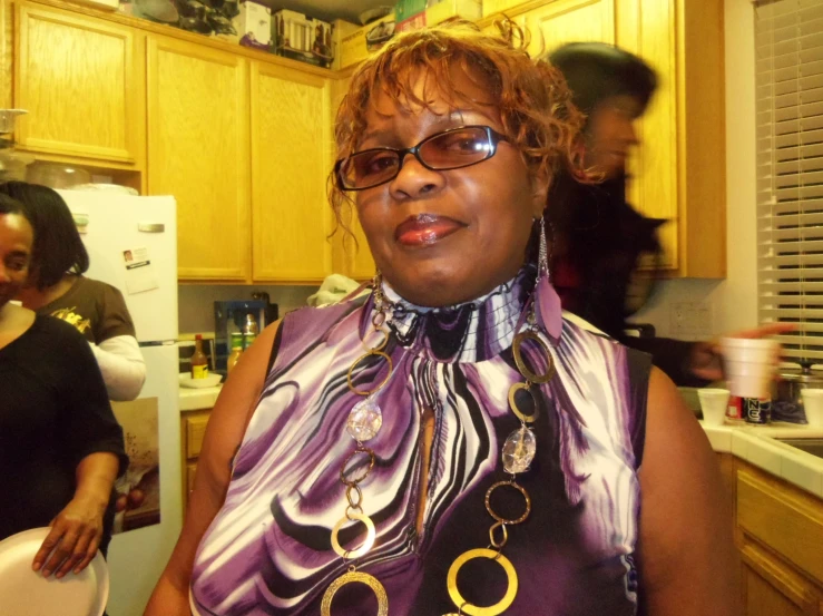 an older woman wearing large gold statement rings and a purple dress in a kitchen