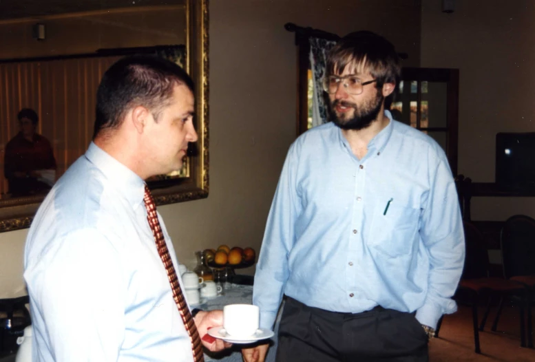 a young man holds a coffee cup and talks to a bearded young man