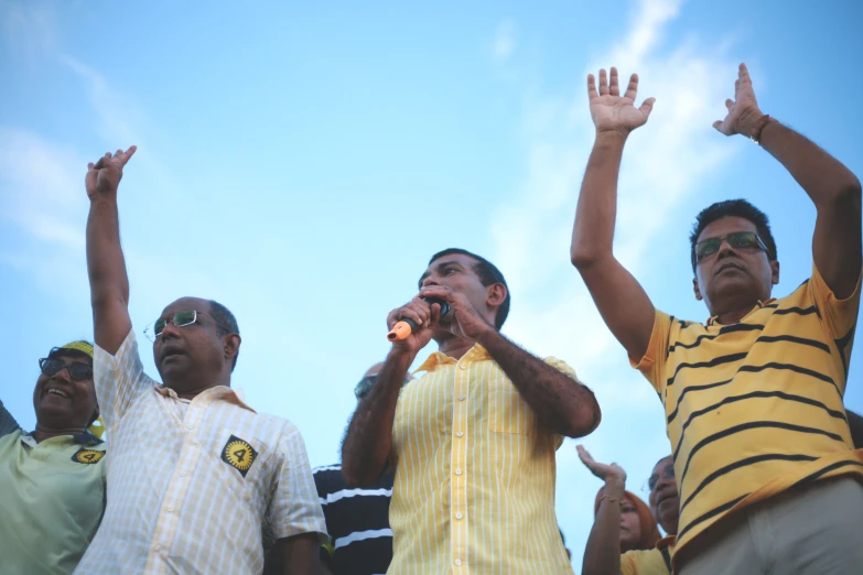 five men, one in a yellow shirt, and two in a black and white striped shirt stand holding their arms in the air
