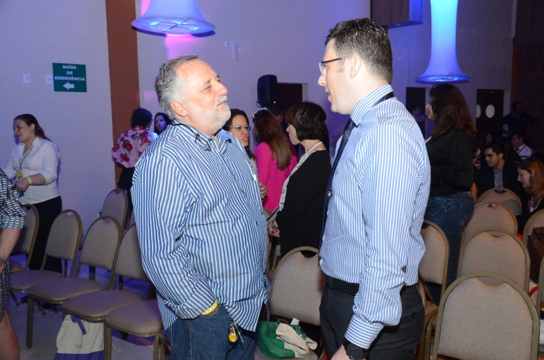 two men talking together in a large room full of people