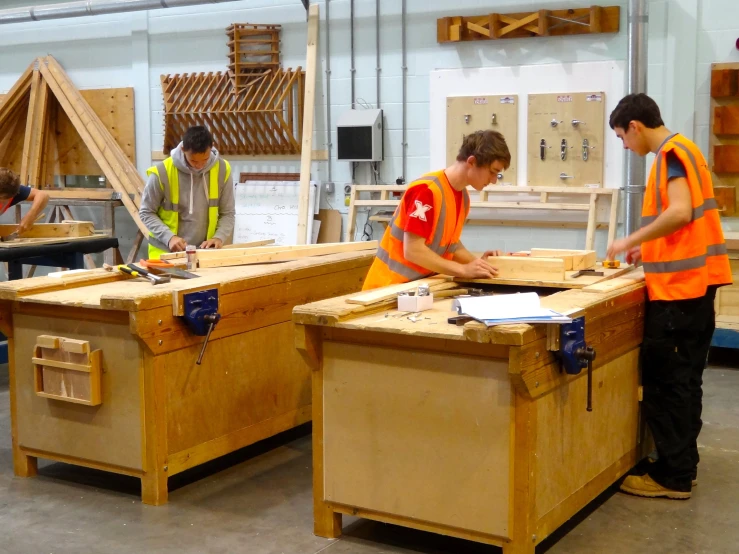 three men in orange vests work on wooden workbenches