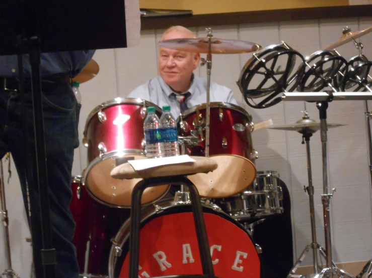 a man with grey hair sitting at a table near some drums