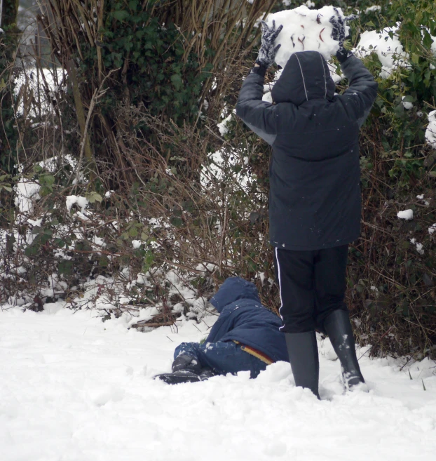 man with black pants and snow - clad jacket standing in the snow while another man lies on ground