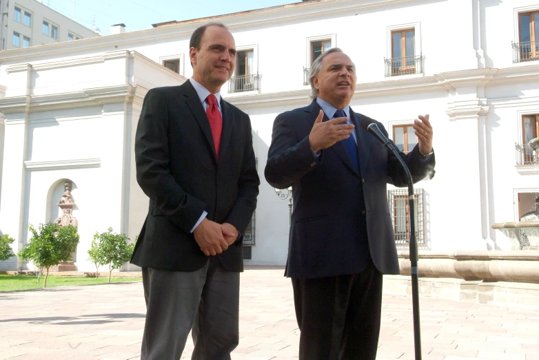 two men standing in front of a building, each holding their hands together