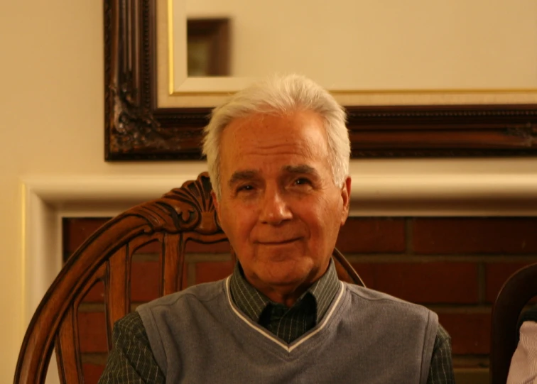 an older man sits in a rocking chair with a brick fireplace and framed artwork above it