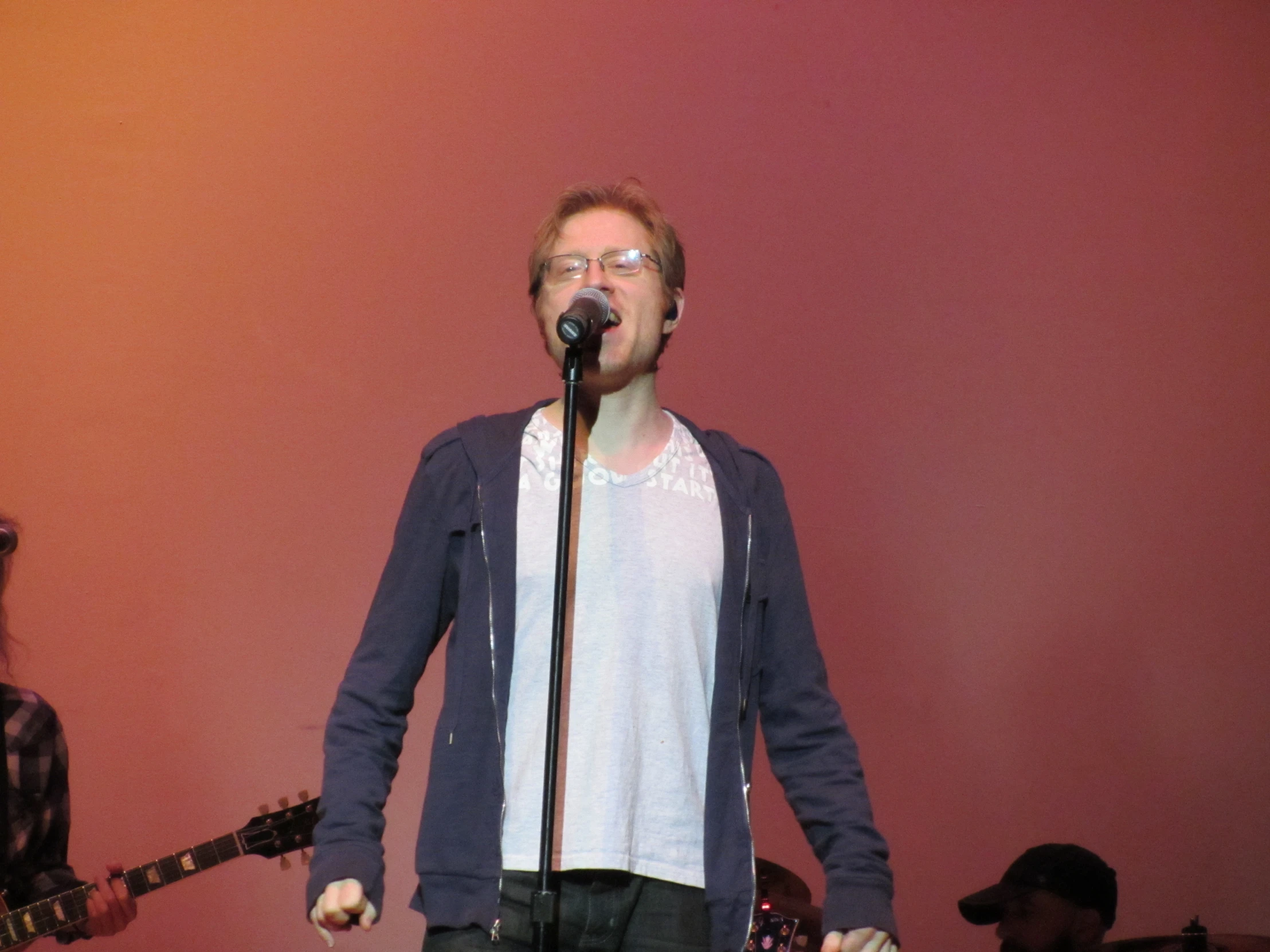 a man with glasses and a white t - shirt sings into a microphone in front of two men