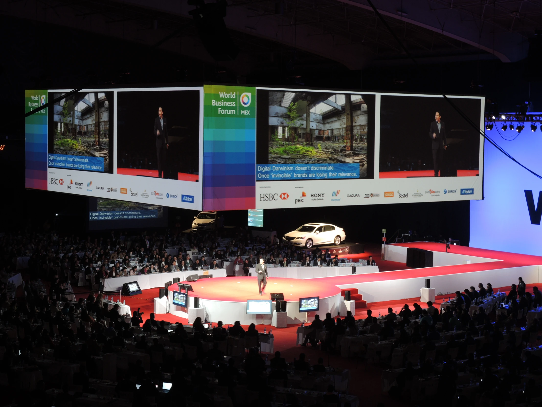 a large auditorium filled with people watching a speaker and a car