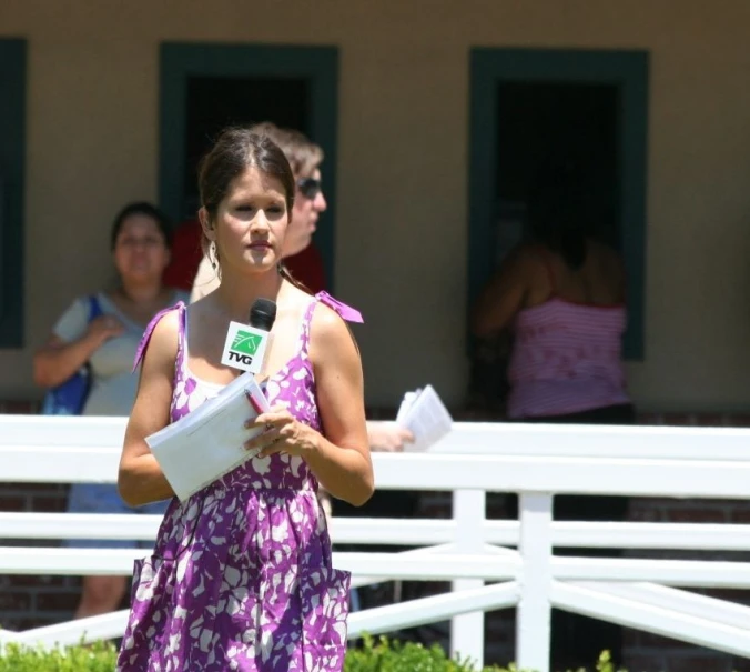 woman in a pink and white floral dress holding a white folder and microphone