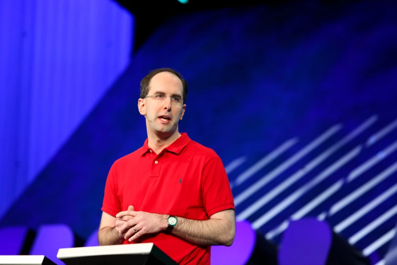 a man in a red shirt stands with his hands clasped on a podium