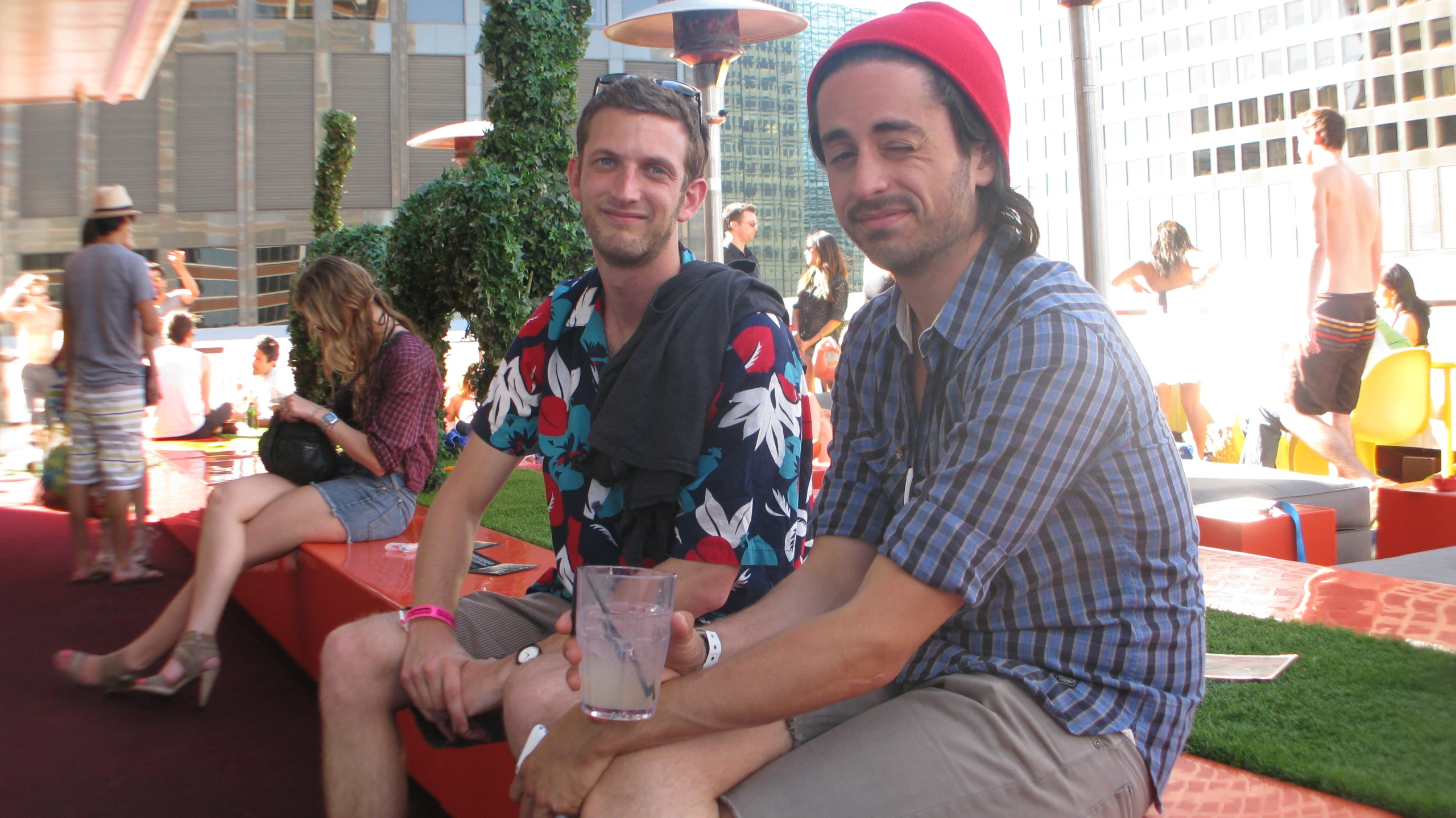 two men are sitting on a bench, one has a drink and the other is standing