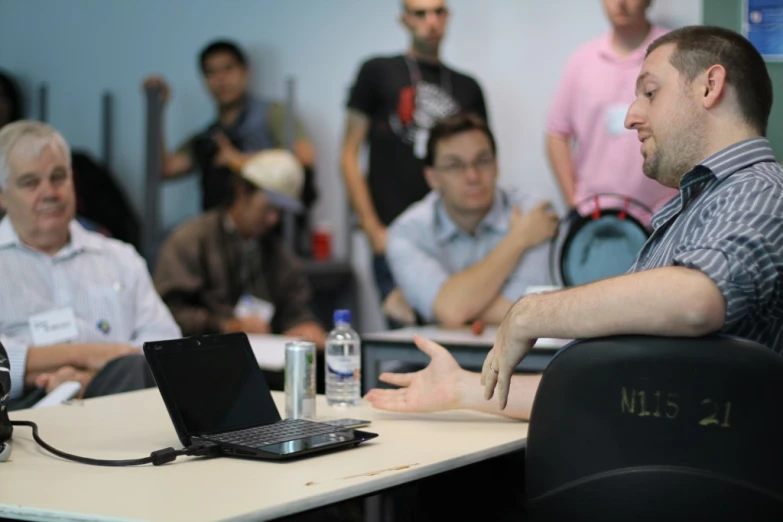 several people sitting at a table listening to a man on his laptop