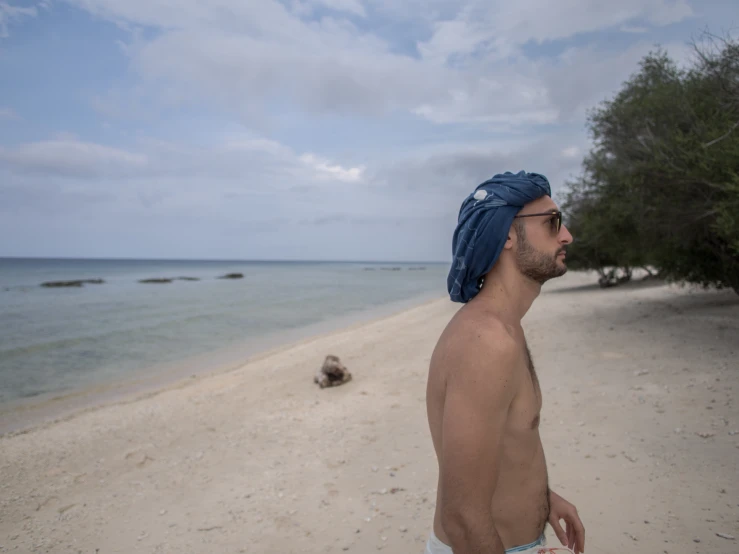 a shirtless man stands on the beach wearing a blue bandana