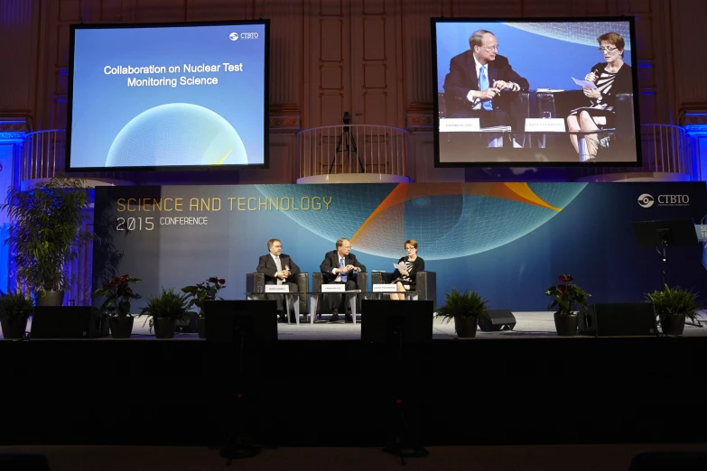 people sit at tables on stage in front of three large screens