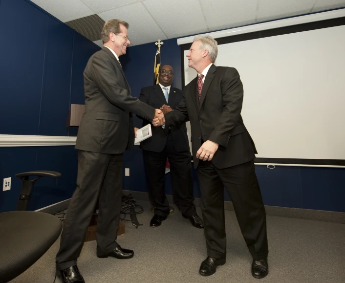 three men in suits shake hands in front of a screen