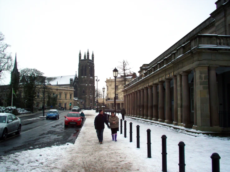 two people walk down a street in winter, with snow on the ground and parked cars