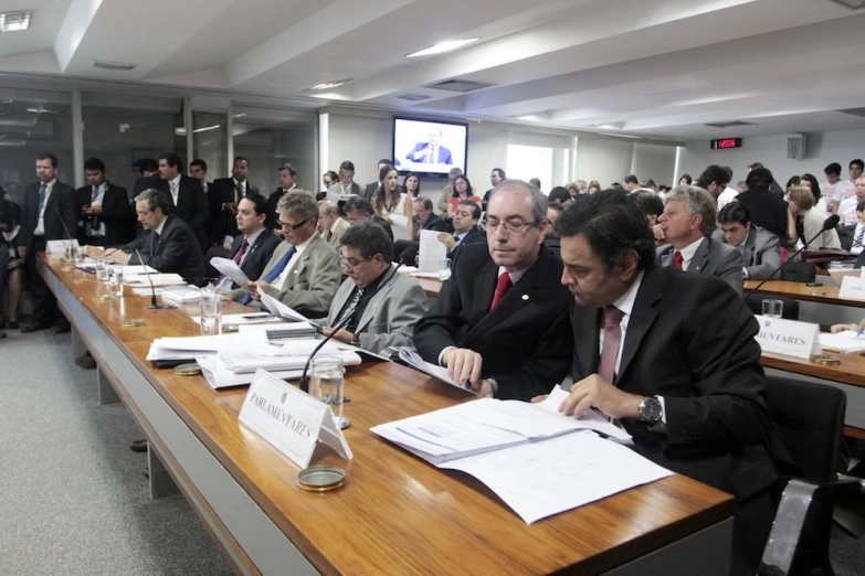 a long table full of people sitting at desks with papers and pen in their hands