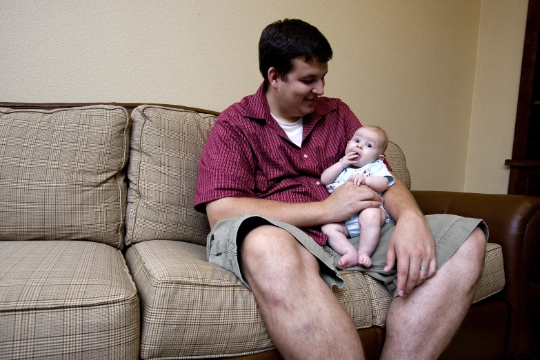 a young man in red shirt sitting on couch holding a baby