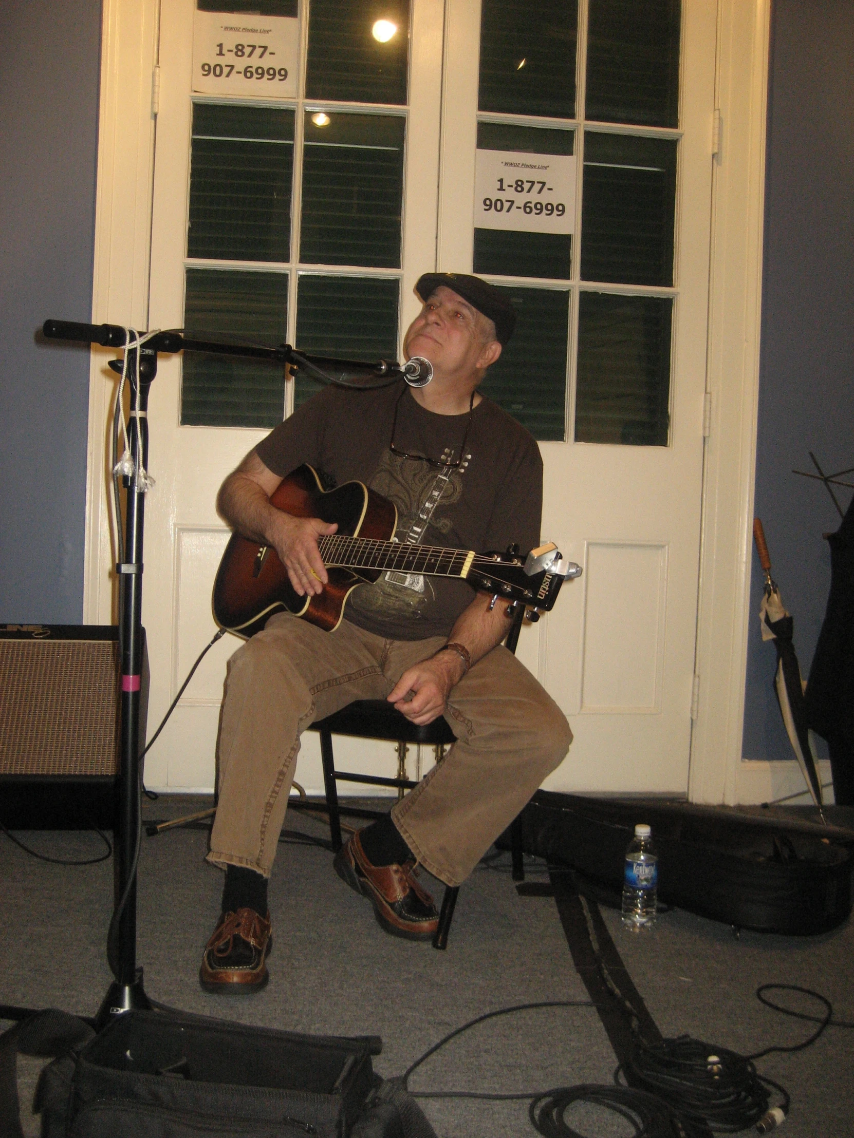 a man in a hat and brown pants sits while playing a guitar