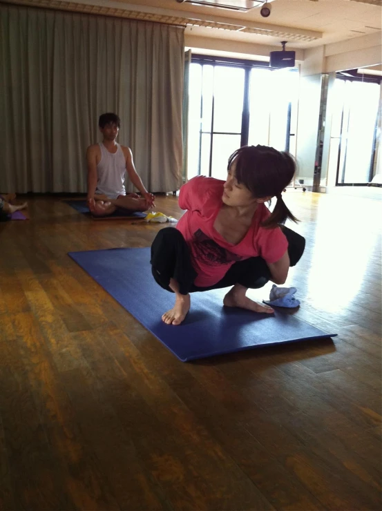 woman standing on the blue yoga mat while another woman sits on the floor
