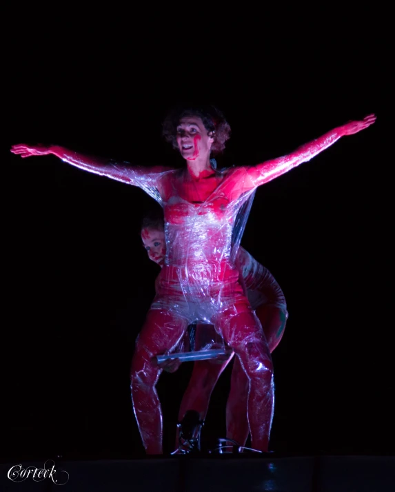 woman in plastic bodysuit standing on stool in dark area with arms outstretched