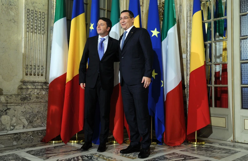 two men standing side by side in front of several flags