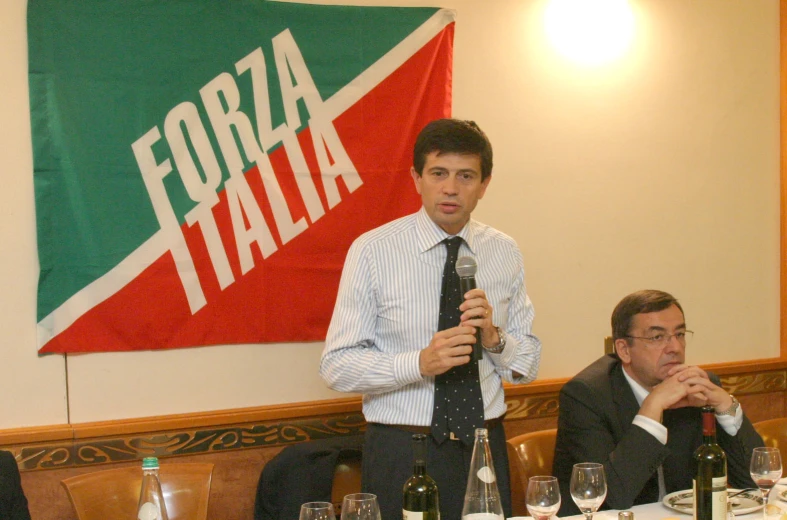 man speaking to several seated men at a long table with a flag behind them