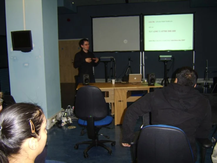 two people are sitting at desks with their backs to the camera while a man stands in front of them