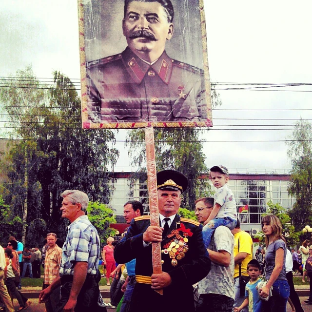 man with hat and cane holding up large poster with image of a dictator