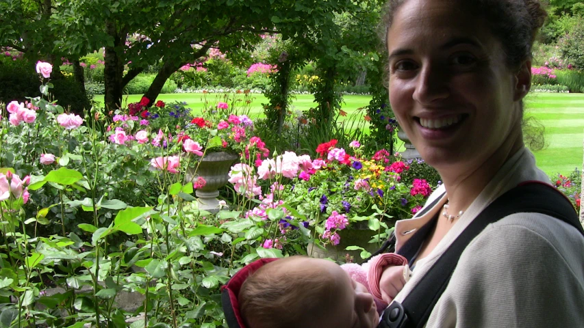 a woman holding a baby in front of a garden full of flowers