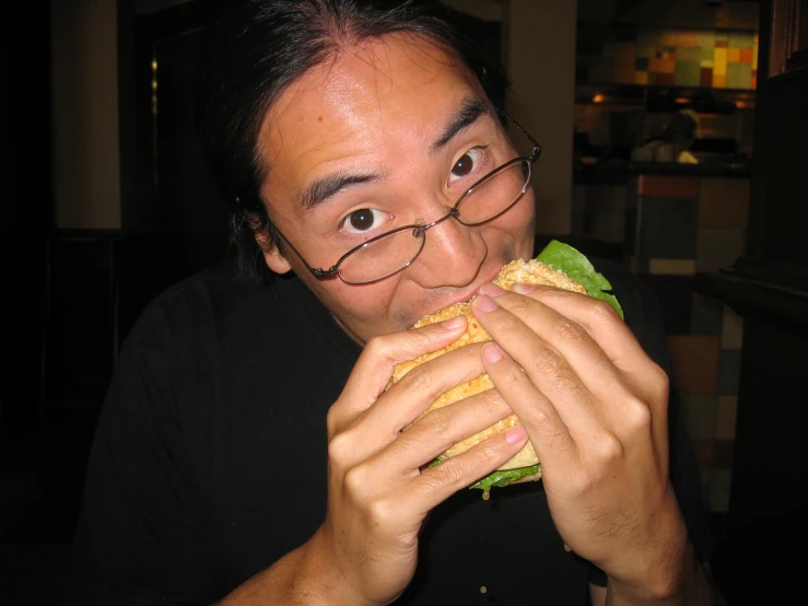 man smiling while eating a big sandwich in a cafe