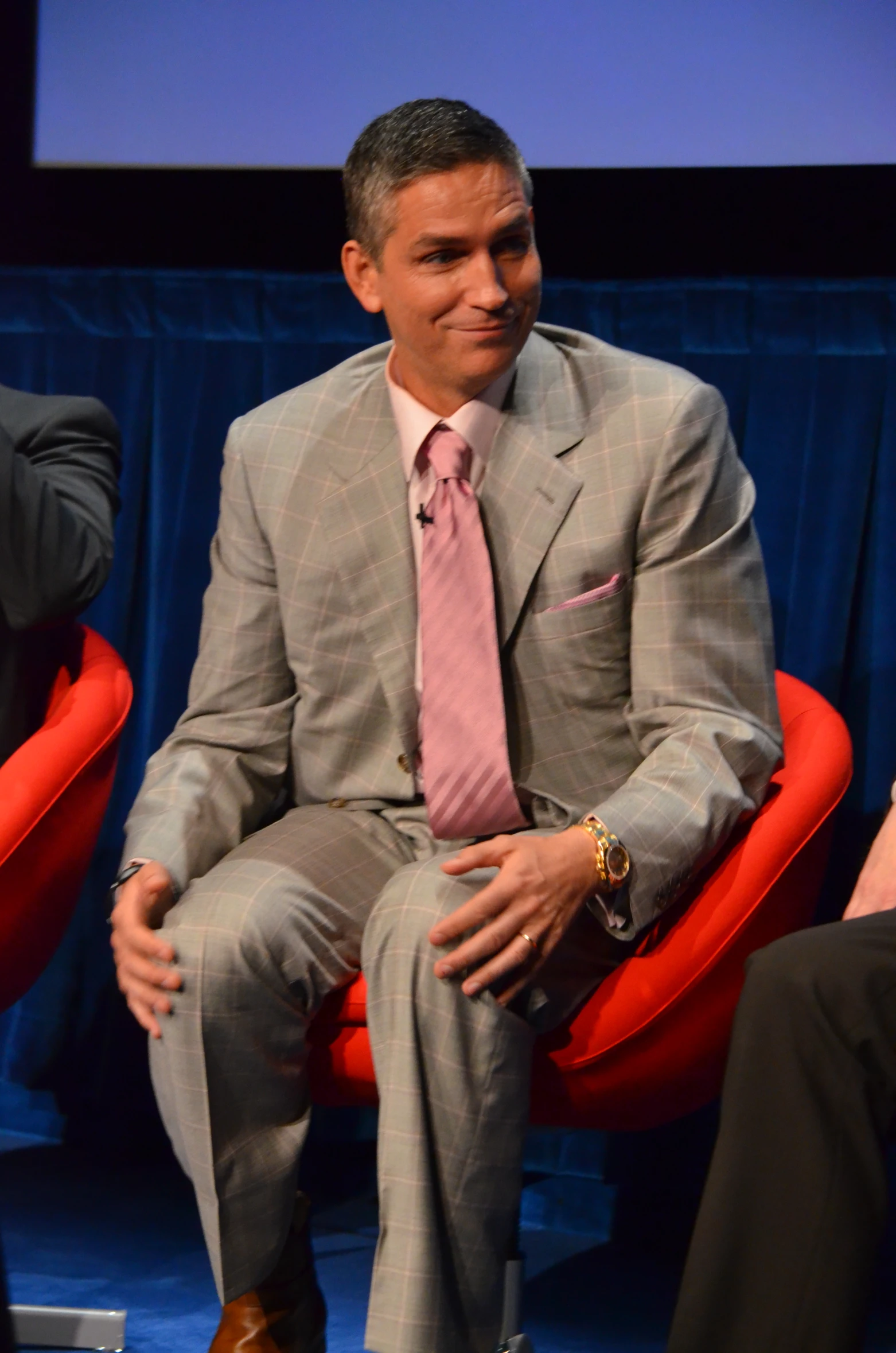 a man in a gray suit and pink tie sitting on red chairs