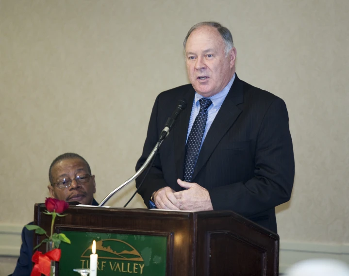 a man stands behind a podium as a third looks on