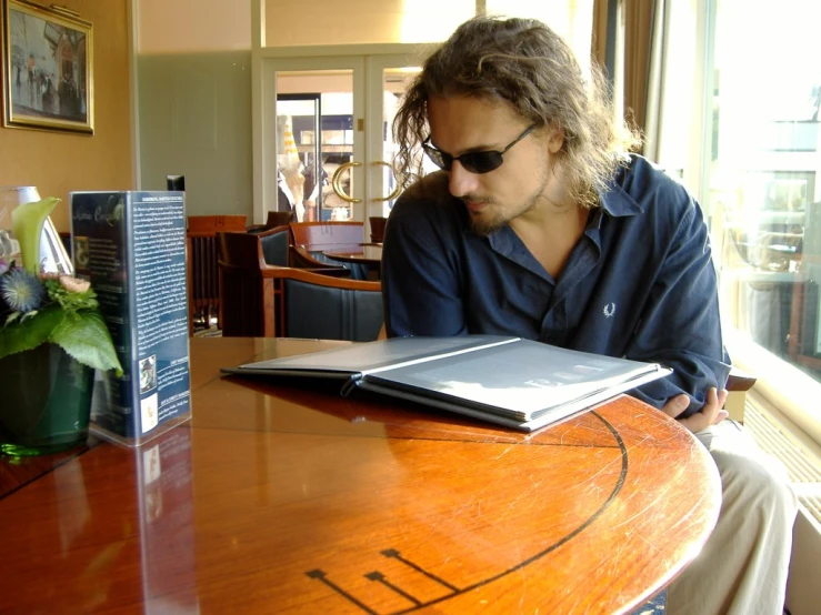 a man wearing sunglasses, sitting at a table with a book