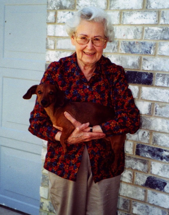 an elderly woman holding a brown baby sheep in front of a building