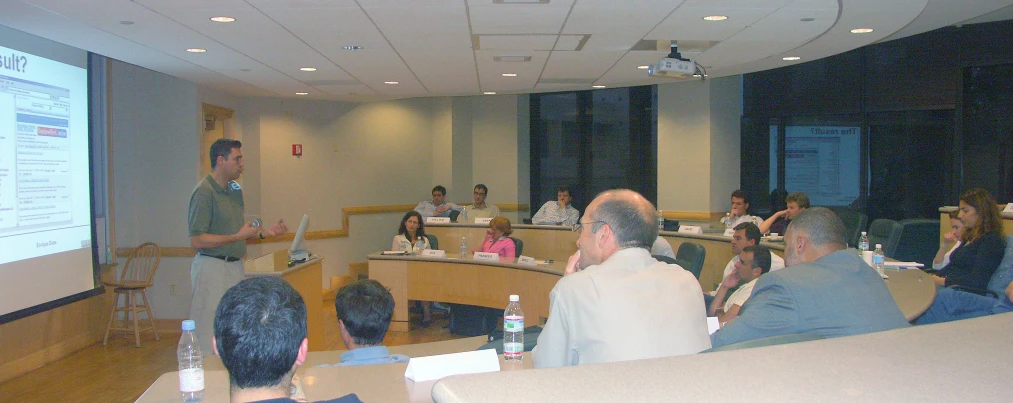 people sitting at tables and in front of a screen listening to someone else talk