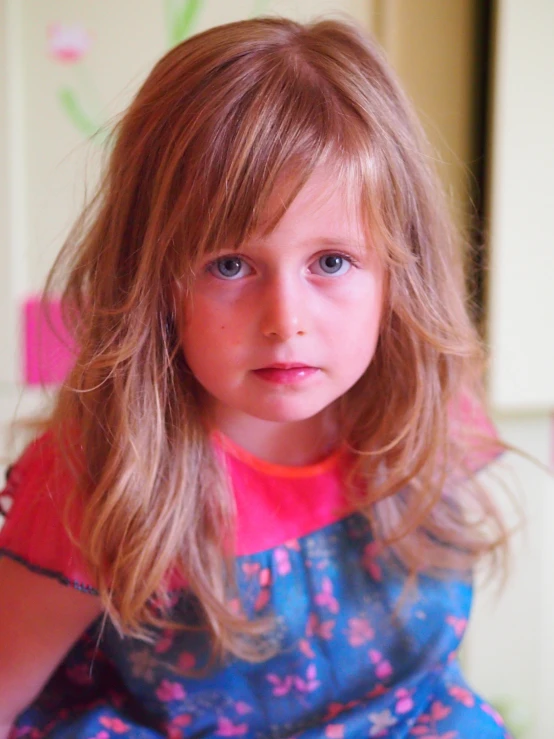 a child with long blonde hair, sits down and looks straight ahead