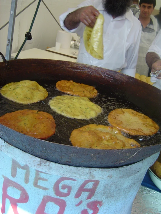 two men in white shirts are preparing pancakes in a large metal pan
