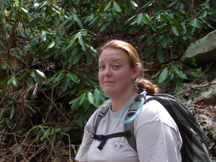 a woman looks at the camera as she stands on the ground in a clearing of bushes and foliage